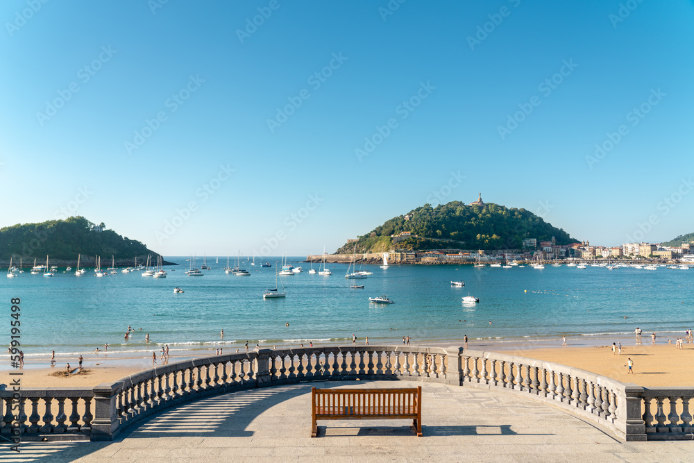 Fototapeta premium SAN SEBASTIAN, Spain July 08 2022: View of isolated bench, on the seafront in San Sebastian. In background is La Concha bay, with yachts docked in de middle and Santa Clara Island. Travel destination