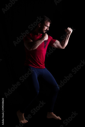 hombre creando una pose de artes marciales con malla azul y camiseta deportiva roja. Fotografía de estudio. Aislado sobre fondo negro. Espacio para texto © Cristina Trujillo