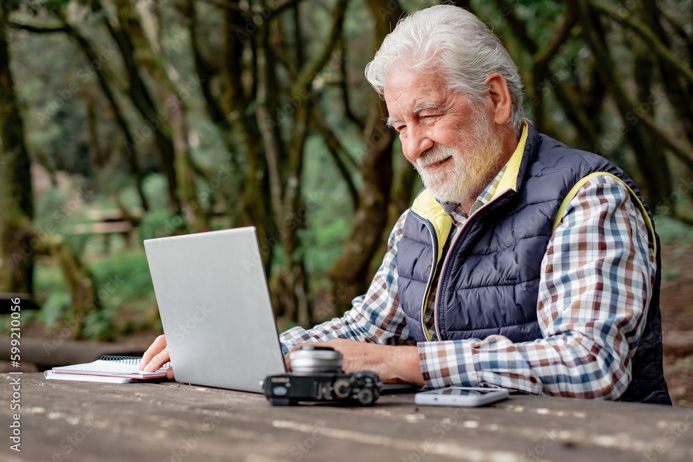 Smiling bearded senior man working on laptop while sitting at a wooden table outdoors in the middle of the park. Concept of smart working in nature, business in fresh air Smiling bearded senior man working on laptop while sitting at a wooden table outdoors in the middle of the park. Concept of smart working in nature, business in fresh air