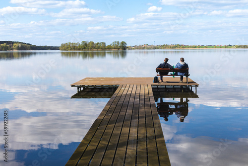 Fototapeta Naklejka Na Ścianę i Meble -  A pair of men, father and son, sitting on a wooden dock by the lake and admiring the view of the water and blue sky