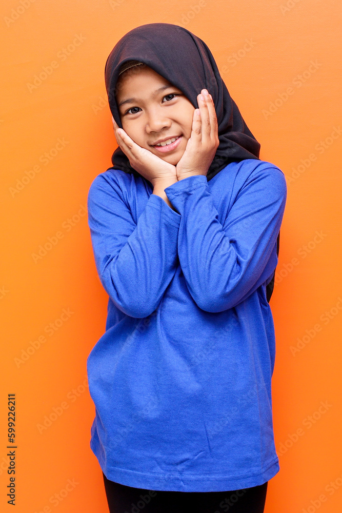 Asian muslim girl wearing hijab with hands on chin, smiling and looking at the camera against orange background. Asian muslim girl wearing hijab with hands on chin, smiling and looking at the camera against orange background.