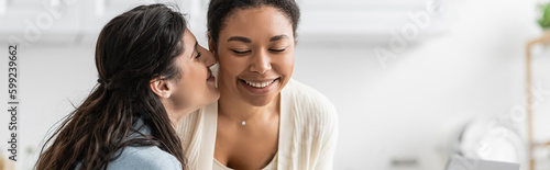 happy lesbian woman kissing cheek of cheerful multiracial girlfriend, banner.