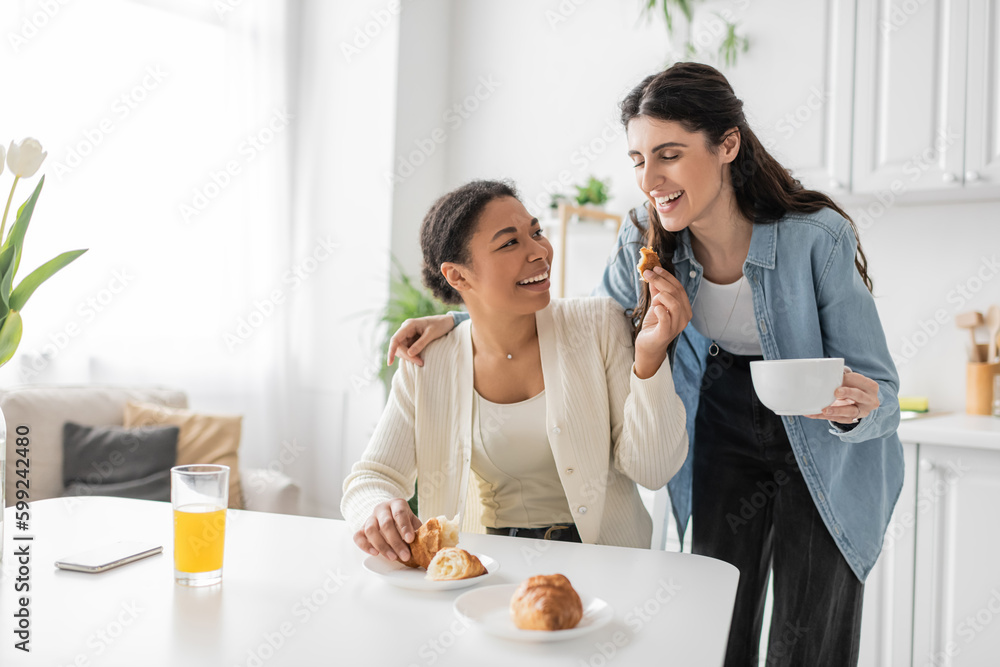 multiracial lesbian woman holding croissant near girlfriend during breakfast in kitchen. multiracial lesbian woman holding croissant near girlfriend during breakfast in kitchen.