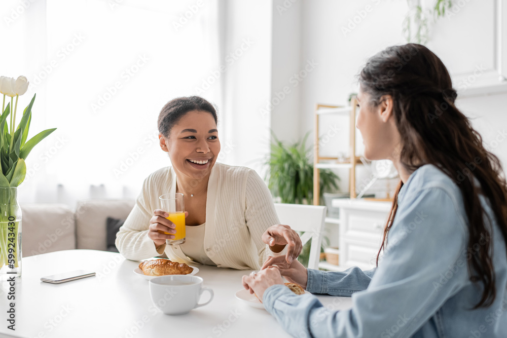 cheerful multiracial lesbian couple smiling while having breakfast in kitchen. cheerful multiracial lesbian couple smiling while having breakfast in kitchen.