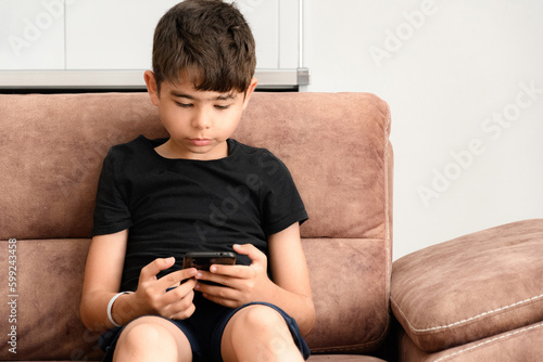 The boy plays on the sofa in the living room. Living room kitchen with sofa. The child plays with gadgets. The boy spends his summer vacation playing games on his phone.
