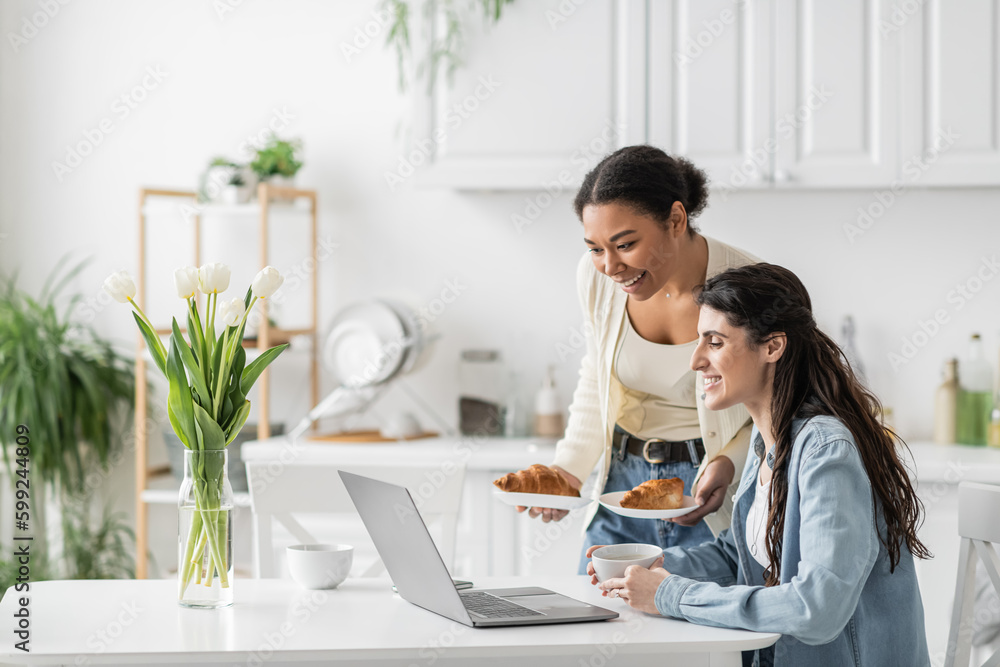 happy multiracial woman holding plates with croissants near girlfriend working on laptop from home.