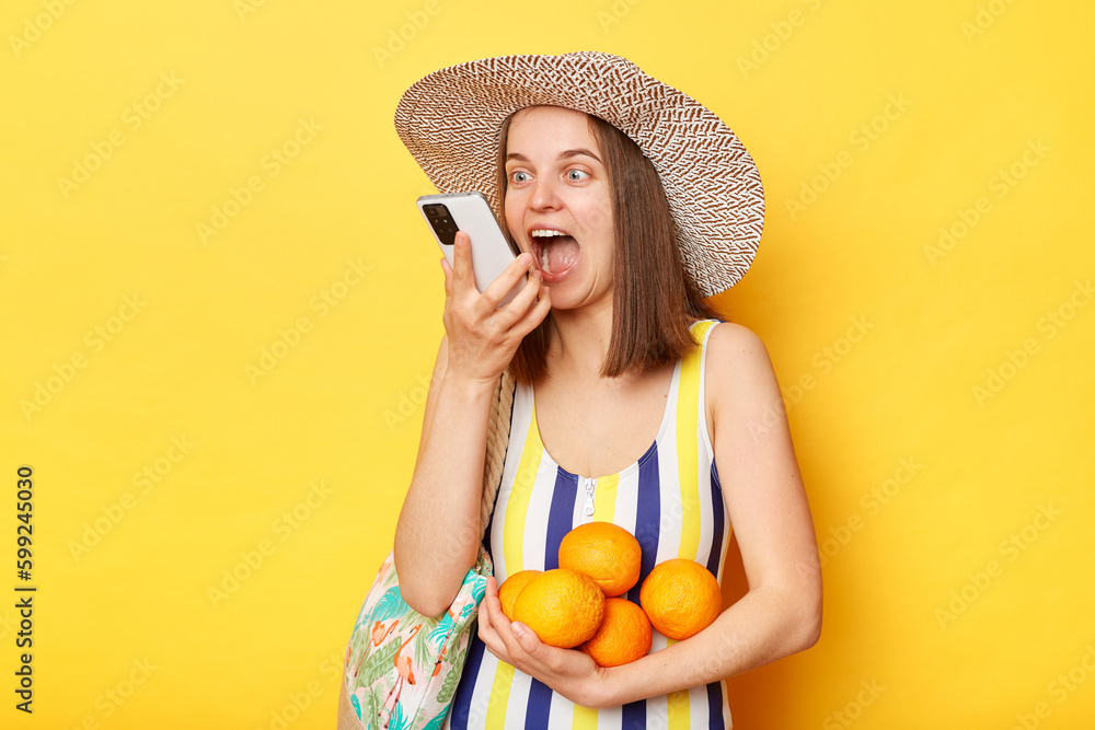 Happy woman wearing striped swimsuit and straw hat isolated yellow background using smart phone recording voice message on summer vacation. Happy woman wearing striped swimsuit and straw hat isolated yellow background using smart phone recording voice message on summer vacation.