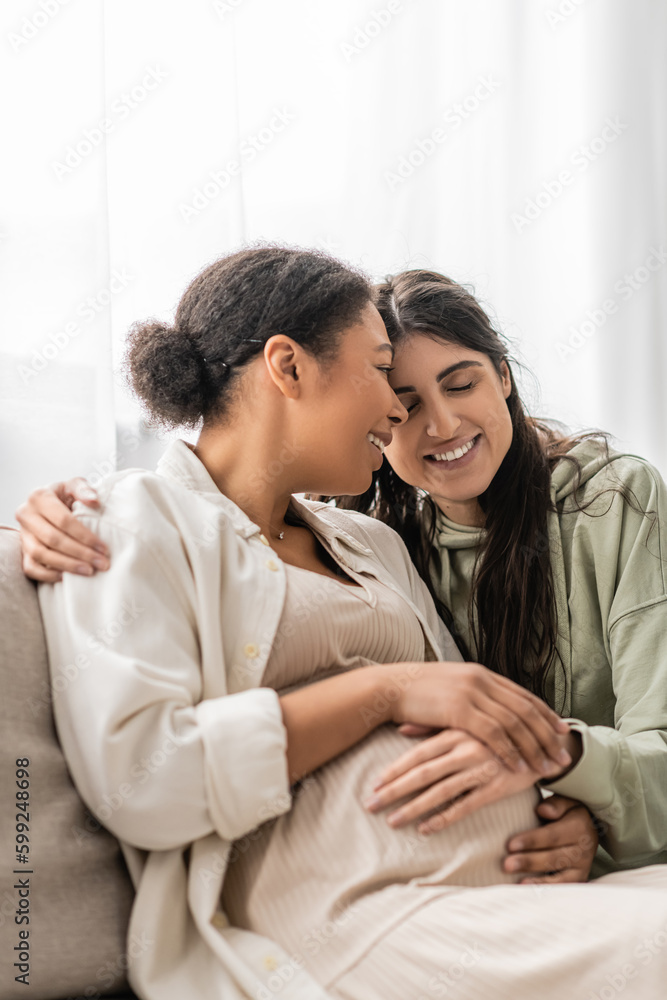 cheerful lesbian woman hugging pregnant multiracial wife while sitting on sofa. cheerful lesbian woman hugging pregnant multiracial wife while sitting on sofa.
