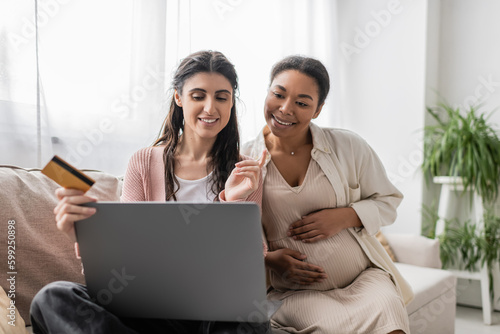 cheerful lesbian woman holding credit card while doing online shopping near pregnant multiracial partner.