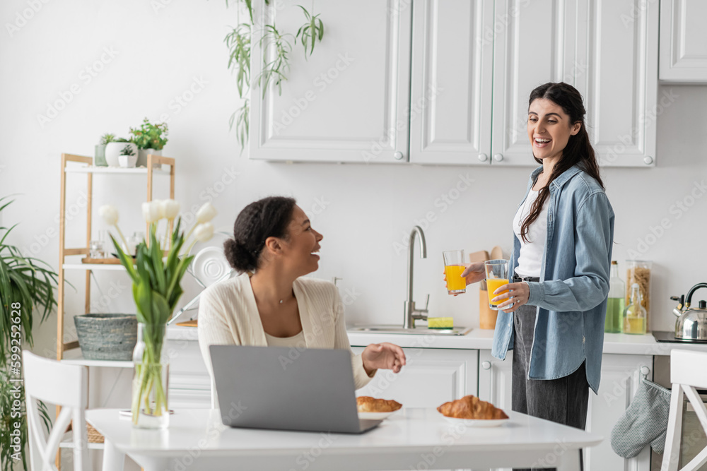 happy lesbian woman holding glasses with orange juice near multiracial girlfriend working on laptop.