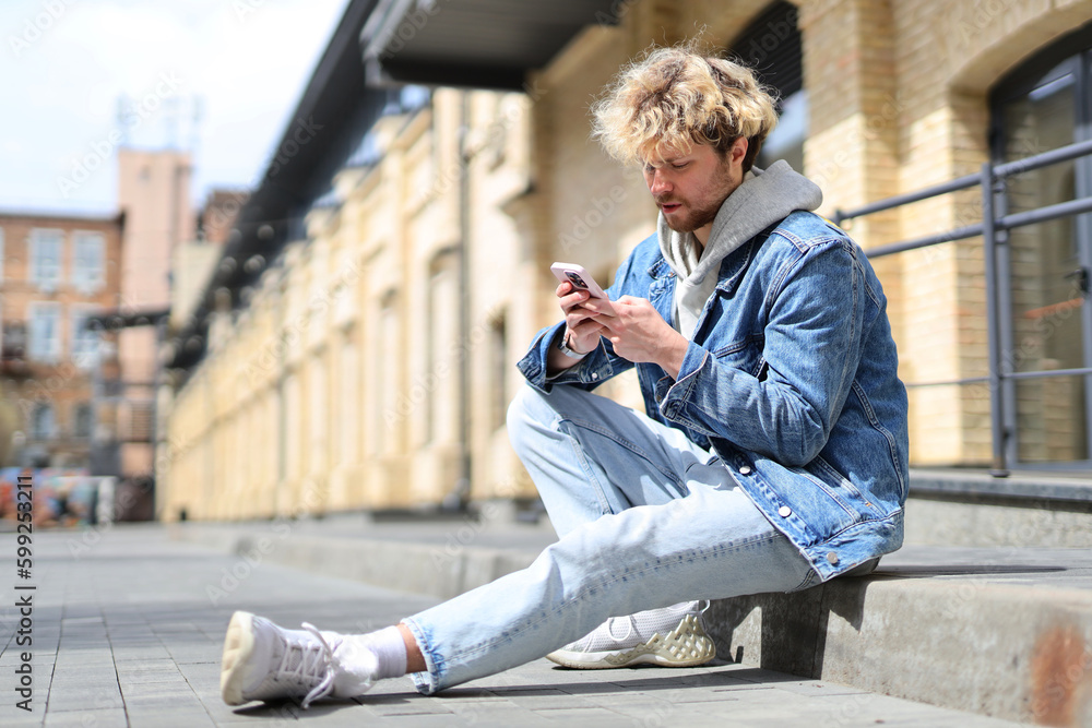 A young man sits outdoors with a smartphone in his hands.