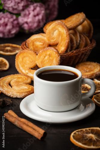 Close-up of cup of coffee with puff pastry, cinnamon stick and lilac flowers on dark wooden table, vertical with copy space