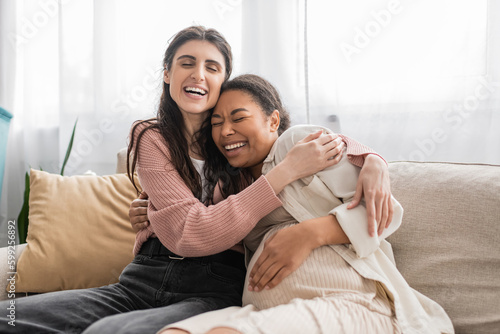 excited lesbian woman hugging pregnant multiracial partner and sitting on couch.