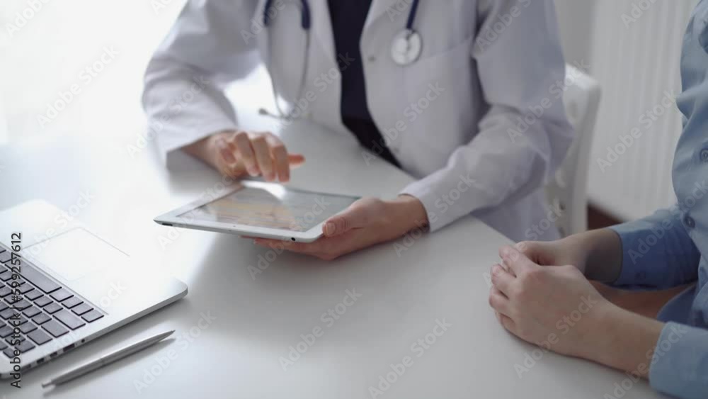 Doctor and patient sitting at the white desk near flair window in clinic. Unknown female physician wearing a white coat uses tablet computer for filling up medical record form. Medicine concept Doctor and patient sitting at the white desk near flair window in clinic. Unknown female physician wearing a white coat uses tablet computer for filling up medical record form. Medicine concept