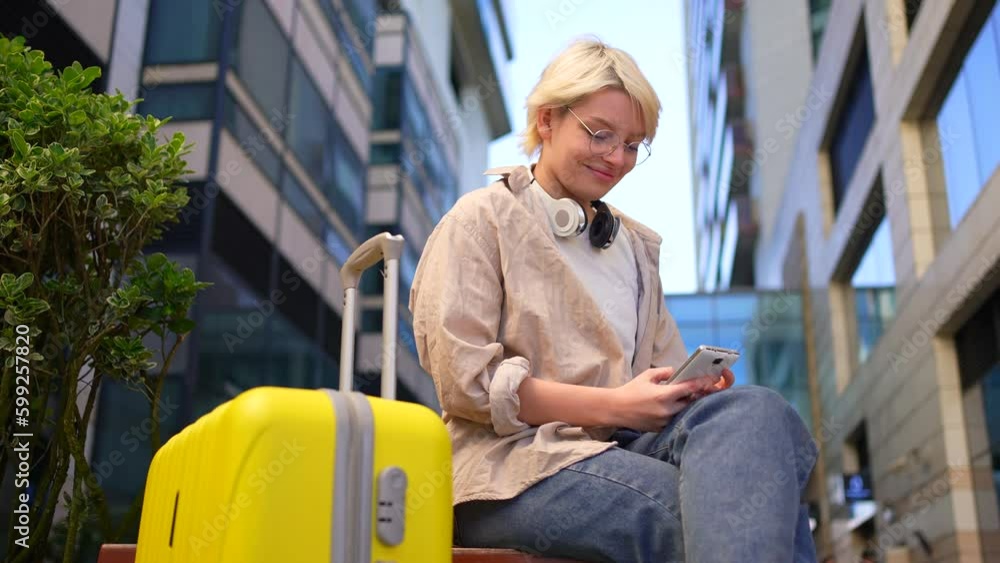 a positive young female traveler on the street with a suitcase is sitting and using a smartphone, smiling a positive young female traveler on the street with a suitcase is sitting and using a smartphone, smiling