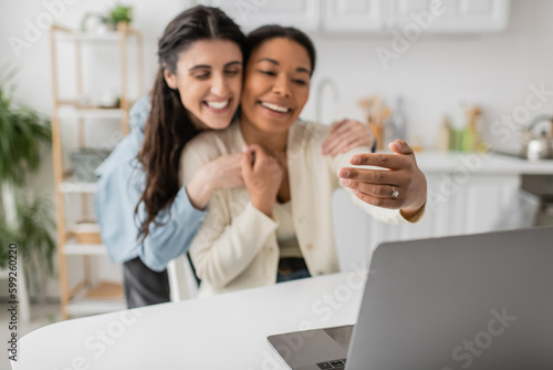 joyful multiracial and lesbian woman showing engagement ring on finger during video call.