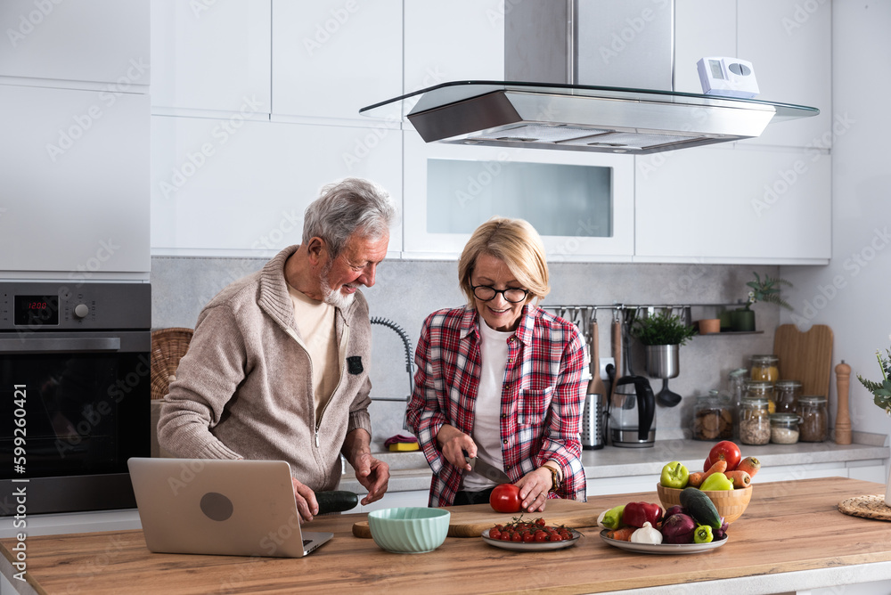 Happy old middle aged senior couple using laptop computer preparing healthy food diet vegetable salad at home together, searching recipes, ordering shopping online, watching cooking class in kitchen. Happy old middle aged senior couple using laptop computer preparing healthy food diet vegetable salad at home together, searching recipes, ordering shopping online, watching cooking class in kitchen.