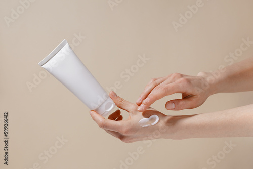 Fragile hands of a woman hold a white mockup tube of facial cream and apply moisturizer to her skin on a beige isolated background. Image for your design.