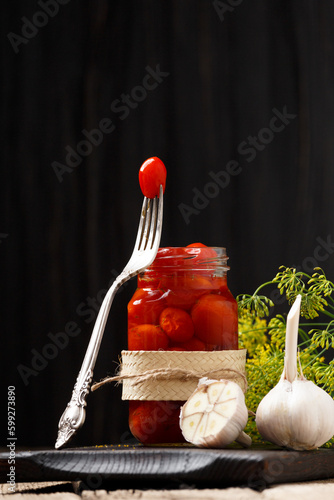 Pickled tomatoes in an open jar, one tomato on a fork, garlic close-up on a dark wooden background.