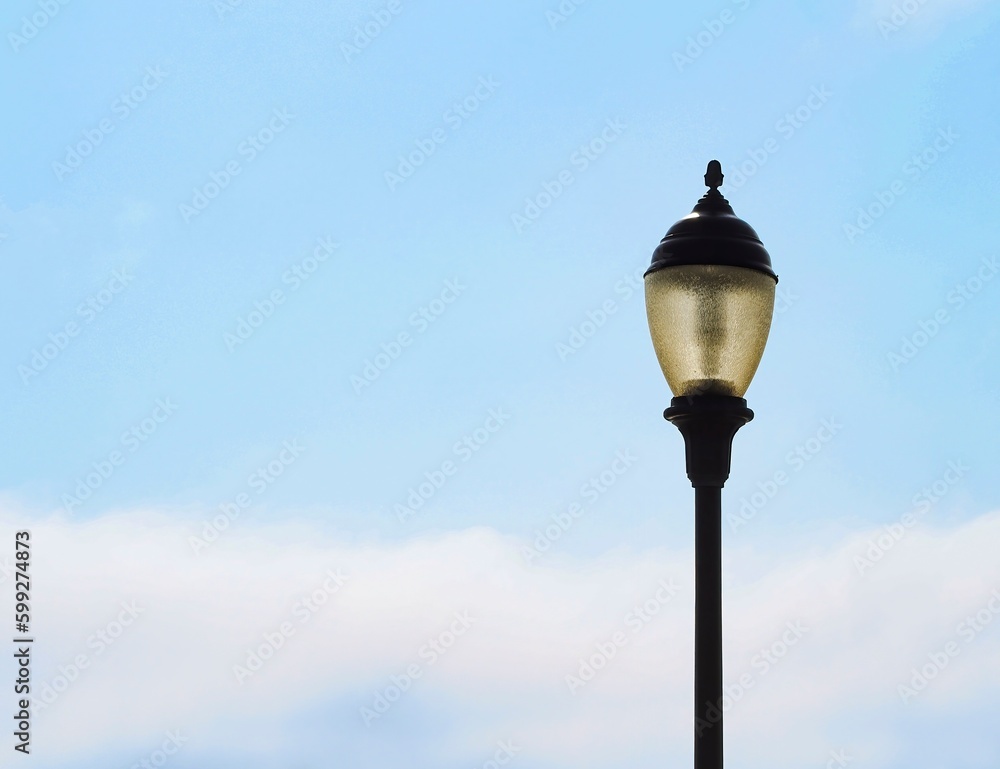 Old-fashioned lamp post, street light, light pole in daytime, with glass lantern, against light blue sky with clouds, copy space.