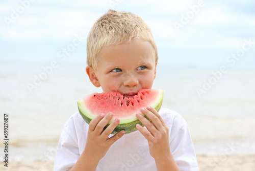 Cute sun tanned blonde kid on the coast, holding juicy slice of the watermelon and eating it.Summertime concept.