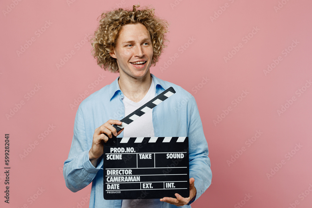 Young caucasian blond man wear blue shirt white t-shirt hold in hand classic black film making clapperboard look aside isolated on plain pastel light pink background studio portrait Lifestyle concept