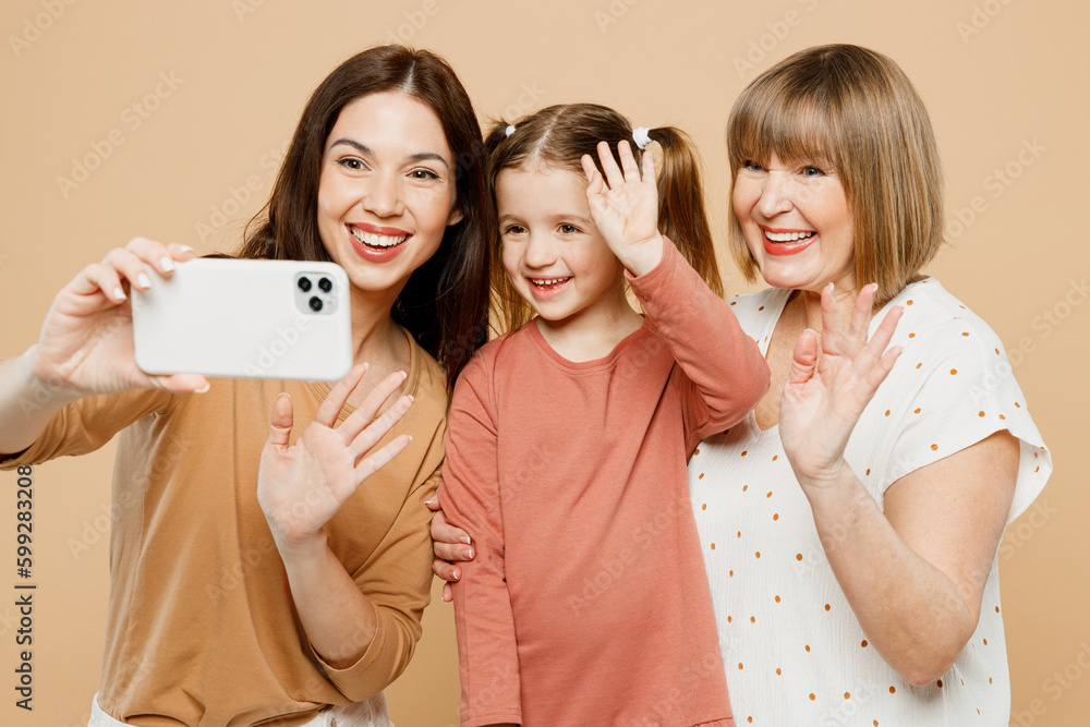 Happy positive women wear casual clothes with child kid girl 6-7 years old. Granny mother daughter doing selfie shot on mobile cell phone isolated on plain beige background. Family parent day concept. Happy positive women wear casual clothes with child kid girl 6-7 years old. Granny mother daughter doing selfie shot on mobile cell phone isolated on plain beige background. Family parent day concept.