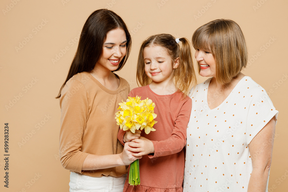 Happy lovely women wear casual clothes with child kid girl 6-7 years old. Granny mother daughter gift giving bouquet of daffodil flowers isolated on plain beige background. Family parent day concept. Happy lovely women wear casual clothes with child kid girl 6-7 years old. Granny mother daughter gift giving bouquet of daffodil flowers isolated on plain beige background. Family parent day concept.