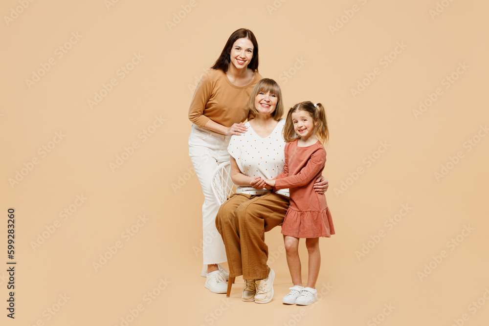 Happy smiling women wearing casual clothes with child kid girl 6-7 years old. Granny mother daughter sit on chair hug look camera posing isolated on plain beige background. Family parent day concept. Happy smiling women wearing casual clothes with child kid girl 6-7 years old. Granny mother daughter sit on chair hug look camera posing isolated on plain beige background. Family parent day concept.