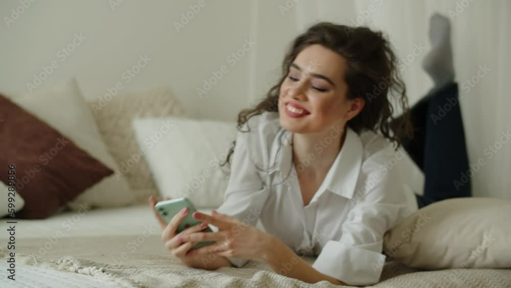 Charming girl is smiling while texting with someone in the bedroom. Woman using smartphone in bed Charming girl is smiling while texting with someone in the bedroom. Woman using smartphone in bed