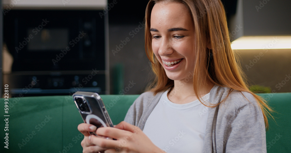 Fototapeta premium Cheerful young woman sitting on a sofa and using smartphone in modern room. Beautiful girl smiling and thinking while chatting on mobile phone.