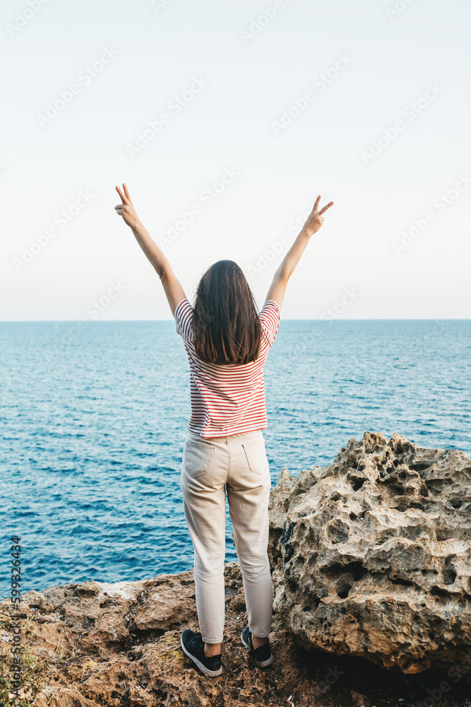 A joyful happy woman, enjoying the beautiful nature of the sea around her.A young woman stands on the seashore and raises her hands up, looking into the distance. 