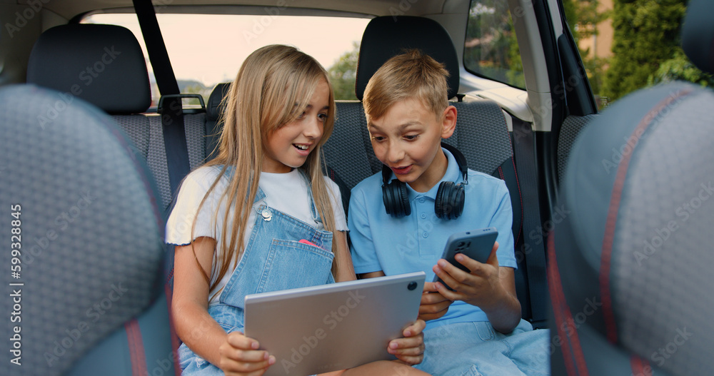 Attractive happy smiling teen kids sitting in the car and applying their tablet pc and phone to review interesting videos Attractive happy smiling teen kids sitting in the car and applying their tablet pc and phone to review interesting videos
