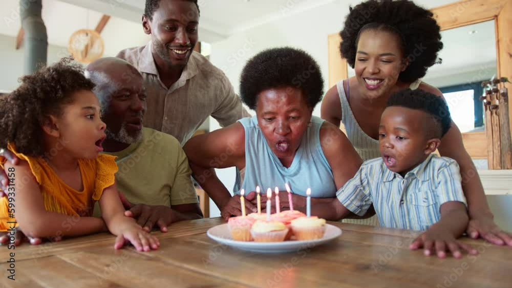 Multi-generation family sitting around table at home celebrating grandmother's birthday singing happy birthday before she blows out candles on birthday cake or cupcakes together- in slow motion Multi-generation family sitting around table at home celebrating grandmother's birthday singing happy birthday before she blows out candles on birthday cake or cupcakes together- in slow motion