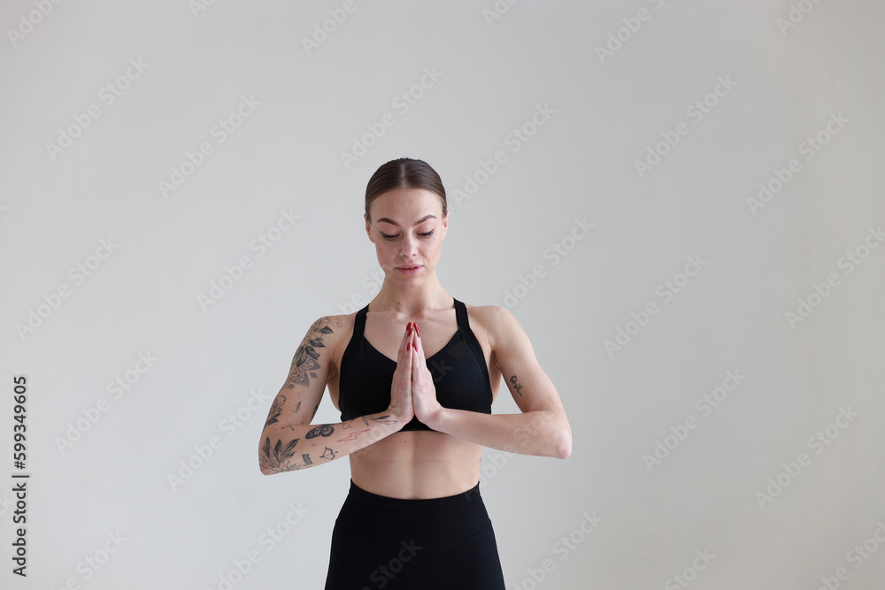 A young girl with brown hair, with a tattoo in a black top and leggings on a white background. Sport, fitness, standing with hands folded A young girl with brown hair, with a tattoo in a black top and leggings on a white background. Sport, fitness, standing with hands folded