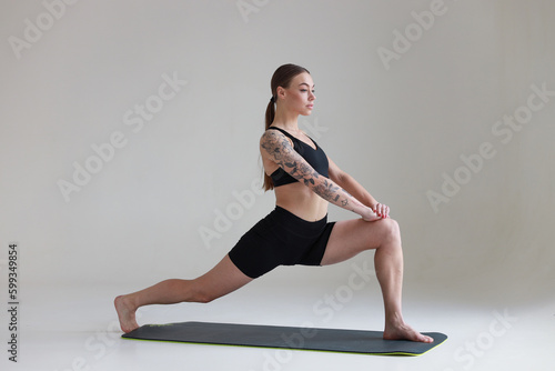 A young girl with brown hair, with a tattoo in a black top and shorts on a white background. Sports, fitness, stretching and yoga