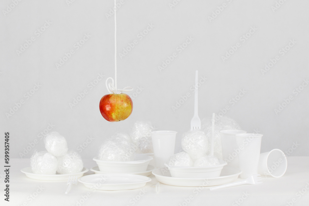 Wiszące na sznurku czerwone jabłko nad plastikowymi jednorazowymi naczyniami  - obrazy, fototapety, plakaty 