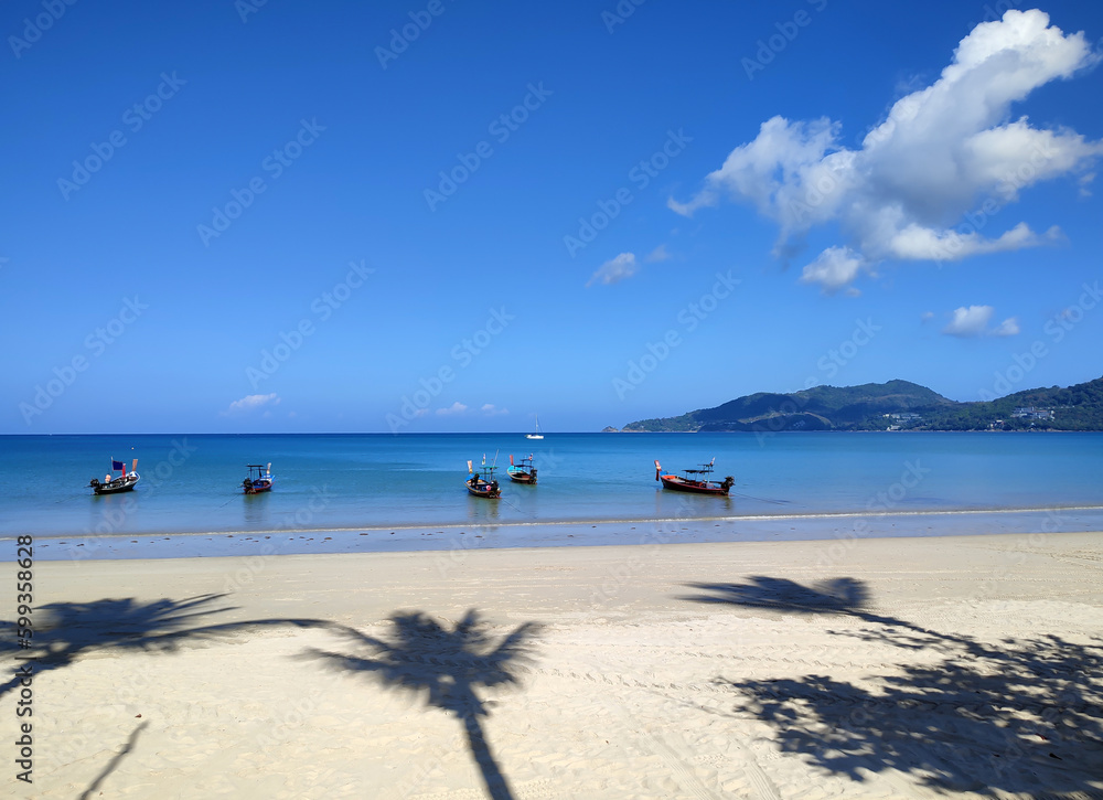 Sandy beach in the tropics, with shadows from palm trees on the sand, overlooking the blue sea, with fishing boats and blue skies