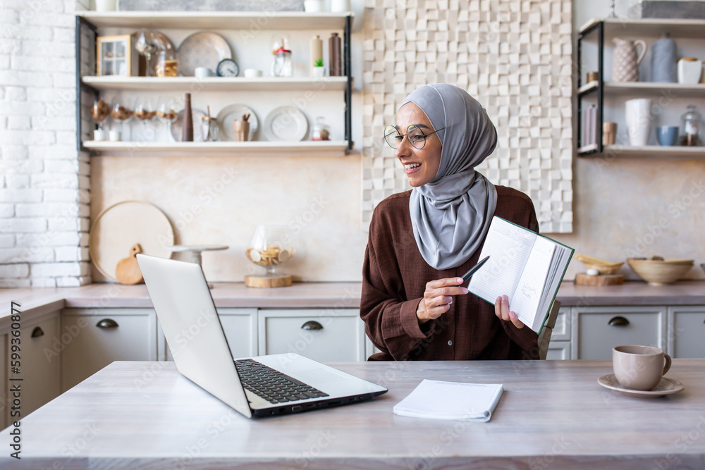 Online training. A young Muslim woman in a hijab teaches at home remotely via zoom from a laptop. Shows the camera a book, a notebook, explains, reads a lecture. Online training. A young Muslim woman in a hijab teaches at home remotely via zoom from a laptop. Shows the camera a book, a notebook, explains, reads a lecture.
