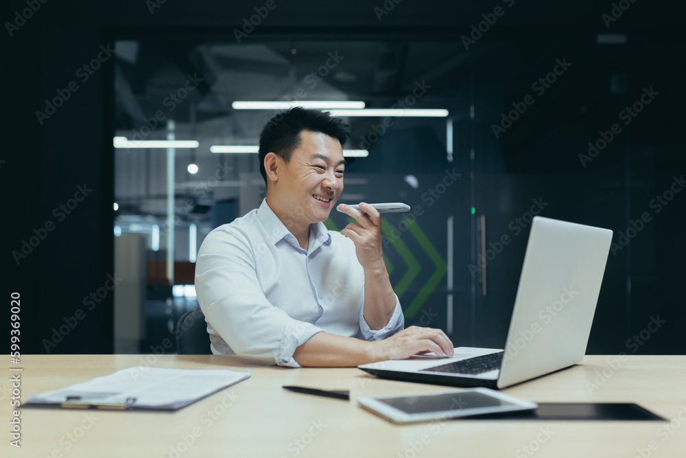 Asian young man businessman working in office with laptop and documents. Talks on the phone through the loudspeaker, records the conversation, smiles.