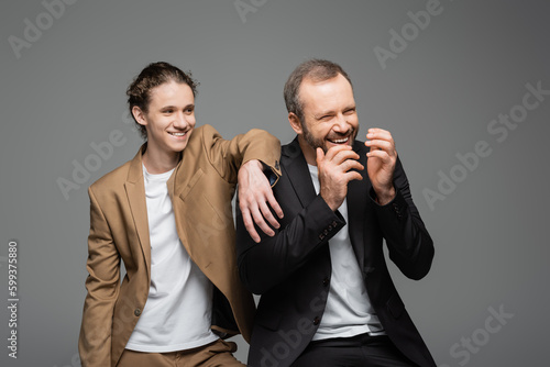 cheerful teenage boy and overjoyed dad in suits laughing isolated on grey.