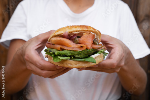 Eating. Closeup view of a woman holding a smoked salmon sandwich with her hands. Delicious layers of bagel bread, pink salmon filet, cream cheese, sliced avocado, capers, onion and arugula.  © Gonzalo