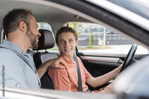 cheerful father putting hand on shoulder of teenage son while teaching him how to drive car. photo