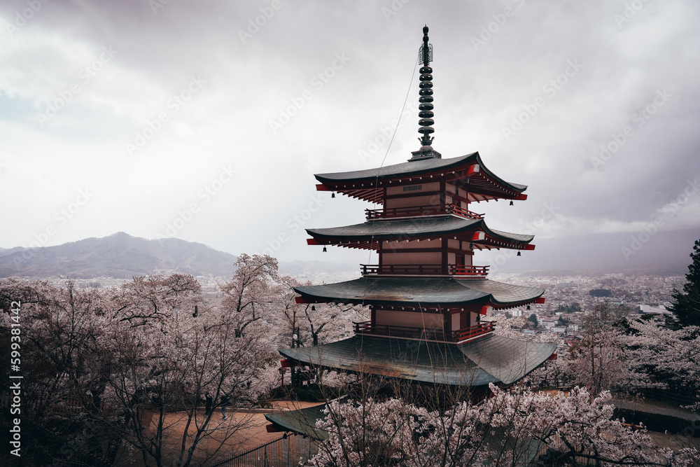 A pagoda towers over a traditional Japanese temple, surrounded by blossoming cherry trees and majestic mountains. The vibrant reds of the plants reflect centuries of faith, culture and history