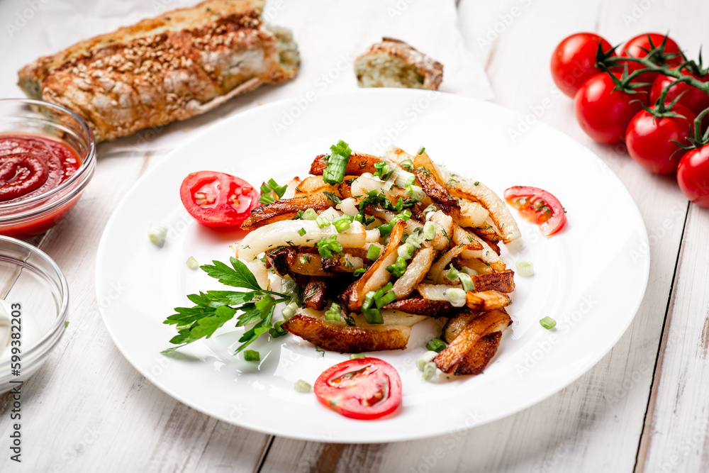 Fried potatoes in a white plate with green onions, parsley, tomatoes on a white wooden background.