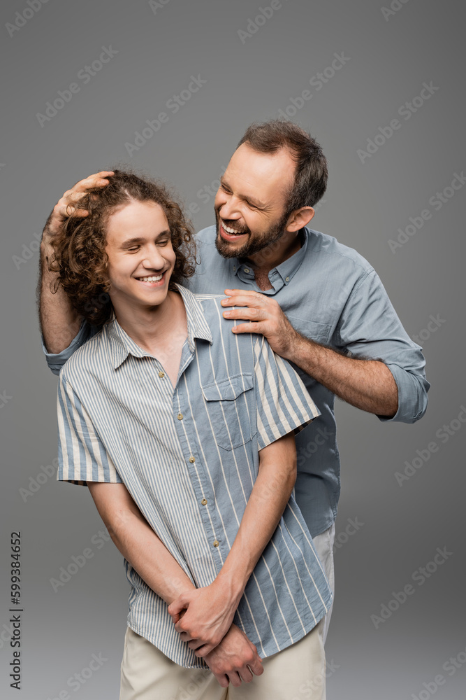 cheerful dad touching curly hair of his teenager son and smiling isolated on grey. cheerful dad touching curly hair of his teenager son and smiling isolated on grey.