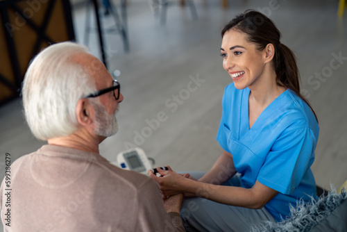 Lovely young nurse smiling and looking at older senior man during home visit check up. Home care, help, support and empathy for older patients.