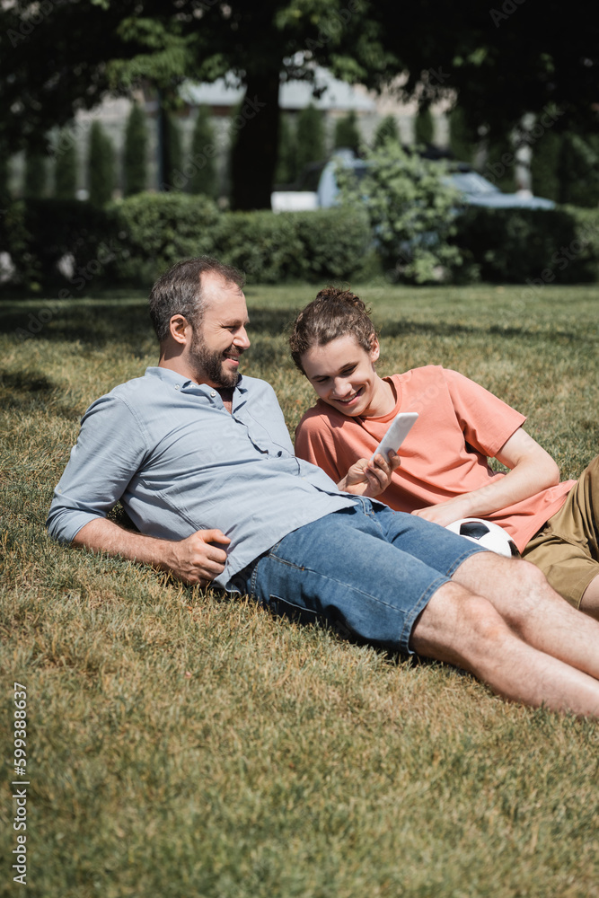 bearded man smiling while using smartphone and resting on lawn near teenage son after playing football. bearded man smiling while using smartphone and resting on lawn near teenage son after playing football.