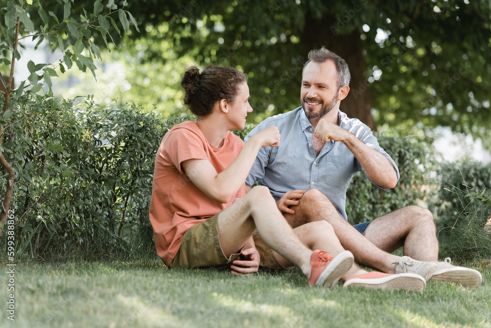 happy father and son doing fist bump while sitting on green lawn in park. happy father and son doing fist bump while sitting on green lawn in park.