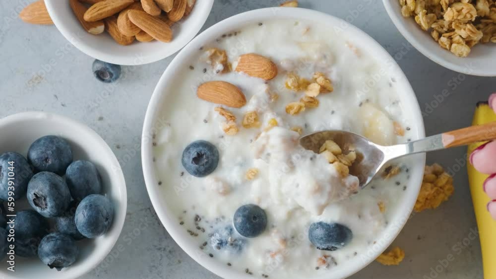 Top view of woman hand eating with a spoon bowl of greek yogurt with oat granola, banana, blueberries and almonds for eating healthy breakfast Top view of woman hand eating with a spoon bowl of greek yogurt with oat granola, banana, blueberries and almonds for eating healthy breakfast